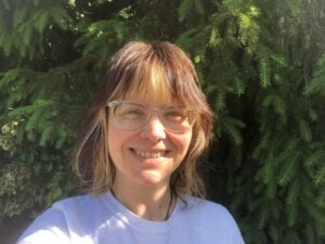 a photo of Kerry smiling in the sun, with a green hedge behind her. She is a white woman with brown hair with a blonde fringe, wearing glasses and a light coloured t shirt.