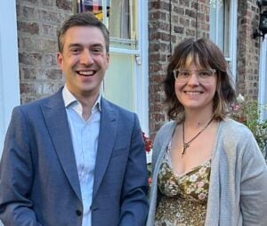 Adrian stands on the left. He is standing outside, wearing a blue suit and a shirt, and smiling. Kerry stands on the right. She is wearing a grey cardigan, a flowery dress and glasses, and is also smiling.