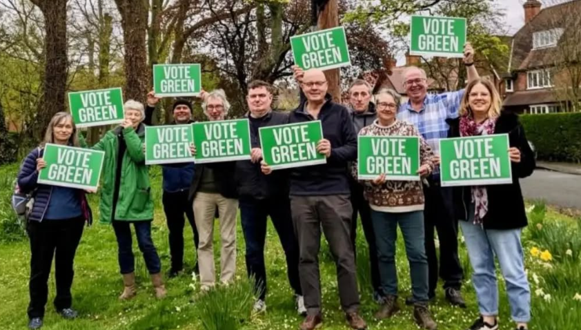 A group of people hold VOTE GREEN signs. They are standing on a green space outside houses. The daffoldils on the edge of the shot hint that it is Spring. 