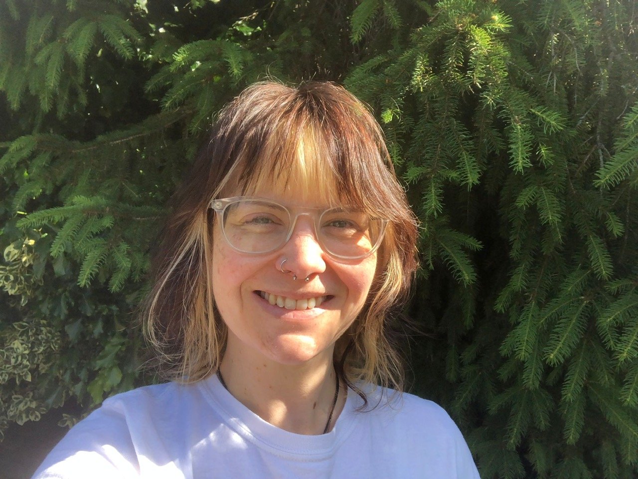 Kerry, a woman with brown hair and blonde highlights, looks into the camera. She stands in front of a green hedge with the sunshine on her face. She is wearing transparent glasses and a white t shirt. 